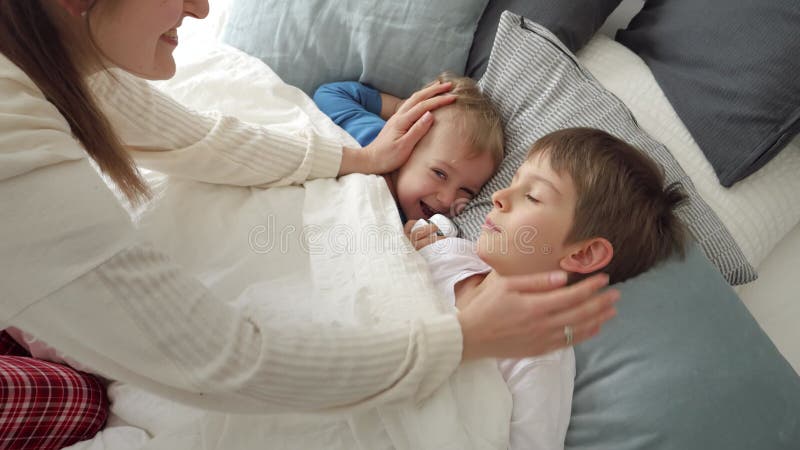 Young caring mother covering her two sons lying in bed with blanket and caressing them. Concept of family happiness.