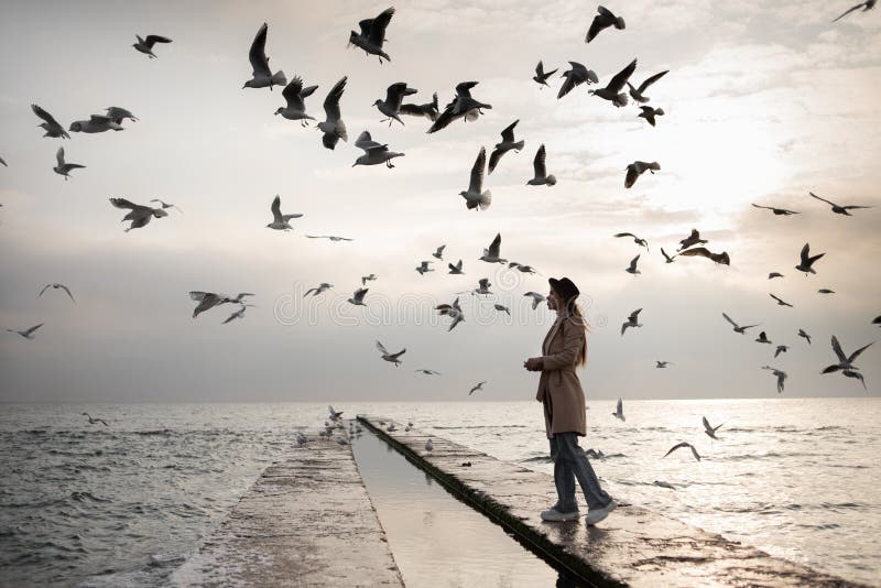 A young Caucasian girl in a stylish coat and hat walks along the pier by the sea and feeds the seagulls. A flock of birds is