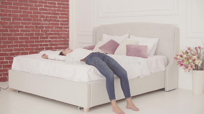 Young Caucasian girl in white shirt and blue jeans falling on the soft white bed. Beautiful woman in casual clothes