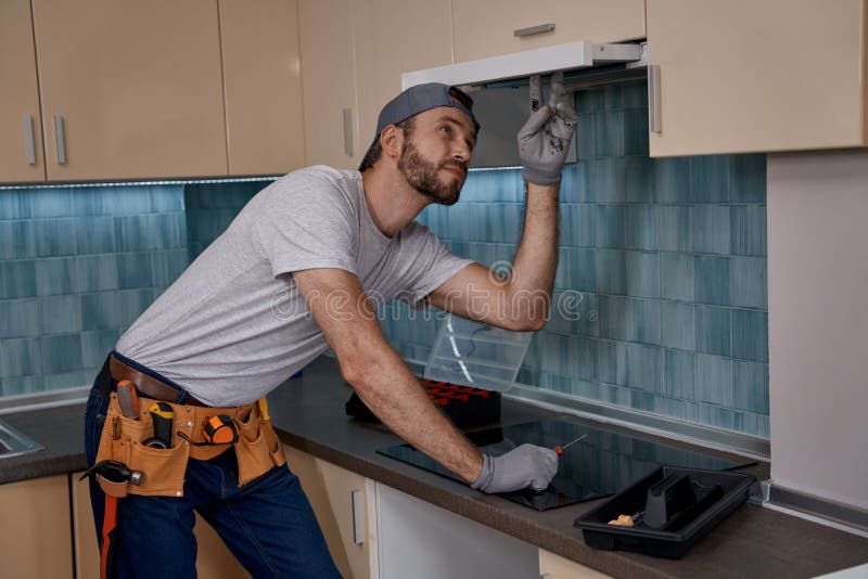 Young caucasian repairman checking work of kitchen hood after installation into new furniture indoors. Home master concept, construction and repair. Young caucasian repairman checking work of kitchen hood after installation into new furniture indoors. Home master concept, construction and repair