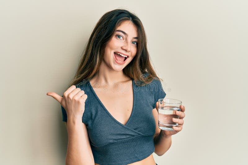 Young caucasian woman drinking glass of water pointing thumb up to the side smiling happy with open mouth