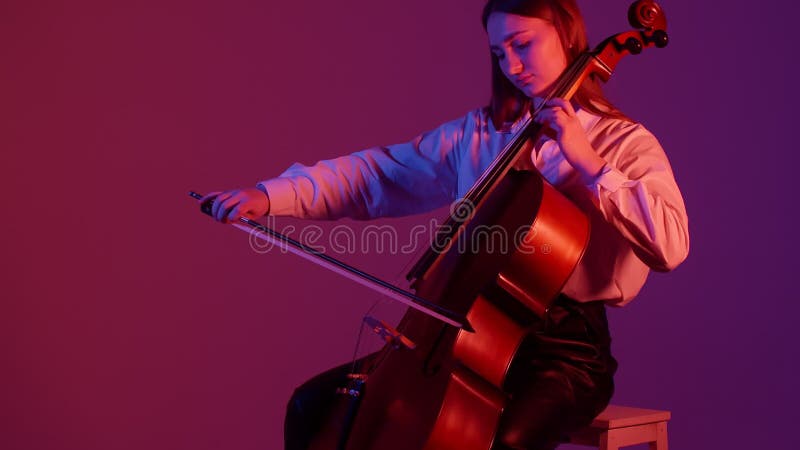 Young Caucasian woman plays the cello in a pink-purple light. Professional cello player plucks strings and plays stock footage