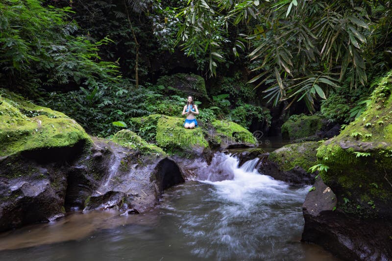 Young Caucasian woman sitting on the rock near the river in Vajrasana or Diamond pose. Hands in namaste mudra. Tropical nature