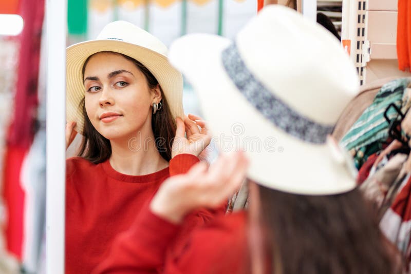 A young Caucasian woman tries on a straw hat for a vacation, and looks in the mirror. The concept of buying clothes stock photo A young Caucasian woman tries on a straw hat for a vacation, and looks in the mirror. The concept of buying clothes stock photo
