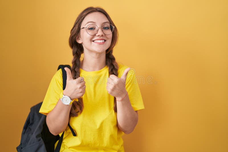 Young caucasian woman wearing student backpack over yellow background success sign doing positive gesture with hand, thumbs up