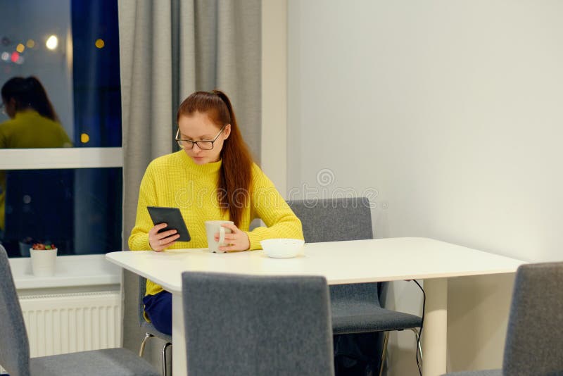 Young caucasian woman in yellow knitted sweater seats at the table in modern light interior and reading an electronic book.