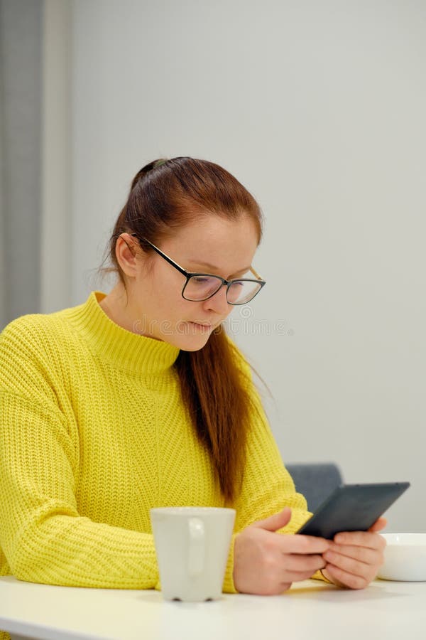 Young caucasian woman in yellow knitted sweater seats at the table in modern light interior and reading an electronic book. Device, hair.