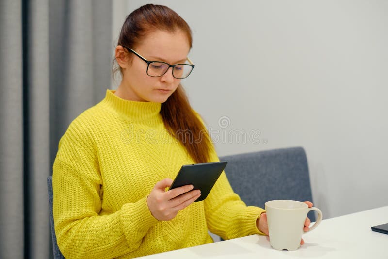 Young caucasian woman in yellow knitted sweater seats at the table in modern light interior and reading an electronic book. Digital, gray.