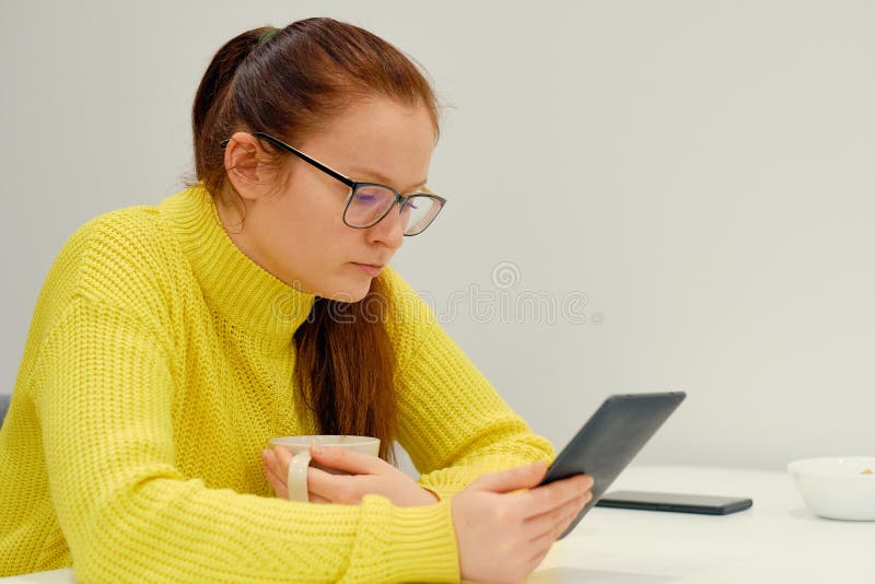 Young Caucasian Woman In Yellow Knitted Sweater Seats At The Table In Modern Light Interior And Reading An Electronic Book.