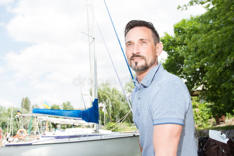 Young cheerful man relaxing on sailboat posing and looking at far on background of boats, sky and trees.