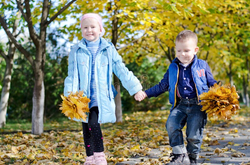 Young children skipping hand in hand while out in the park collecting colourful yellow autumn leaves together