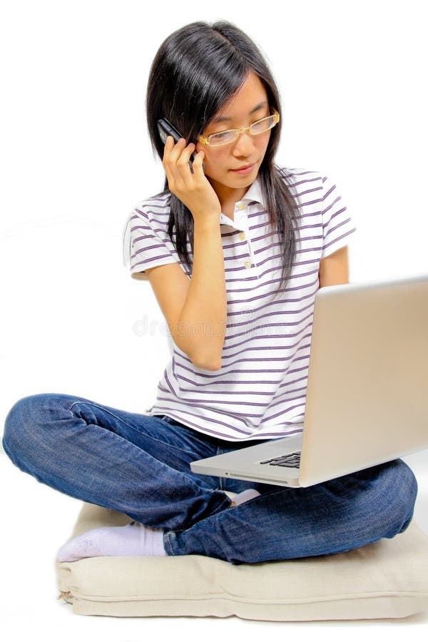 Young chinese woman talking in mobile phone. Sitting on a pillow on the floor. Laptop in front of her