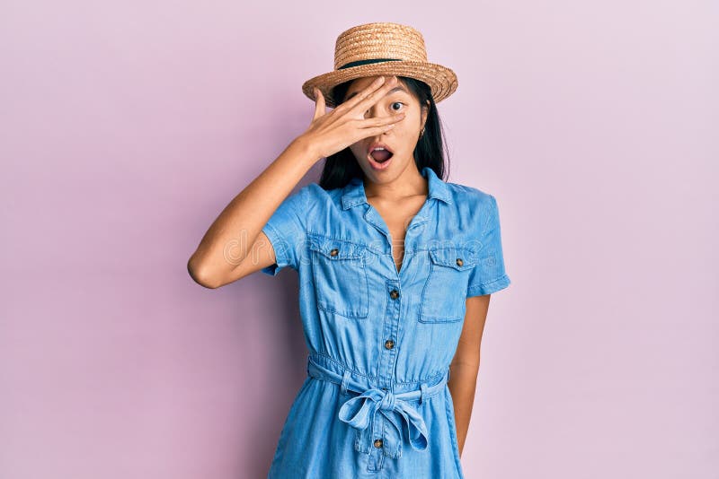 Young chinese woman wearing summer hat peeking in shock covering face and eyes with hand, looking through fingers afraid