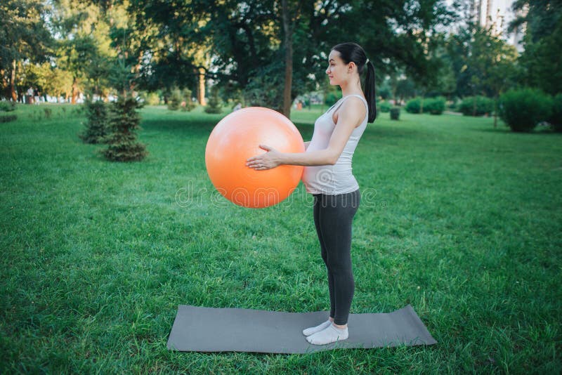 Young concentrated pregnant woman stand on yoga mate in park and hold orange fitness ball. She exercising. stock photo Young concentrated pregnant woman stand on yoga mate in park and hold orange fitness ball. She exercising. stock photo