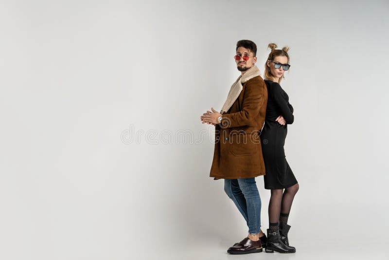 Young couple of bearded men in coat and pretty women in black dress with long blonde hair, both with sunglasses posing in studio on gray background, copy space. Young couple of bearded men in coat and pretty women in black dress with long blonde hair, both with sunglasses posing in studio on gray background, copy space