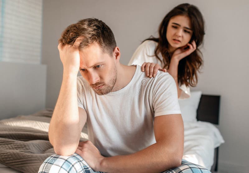 Depressed man is sitting on the edge of bed while his girlfriend is tryin.....