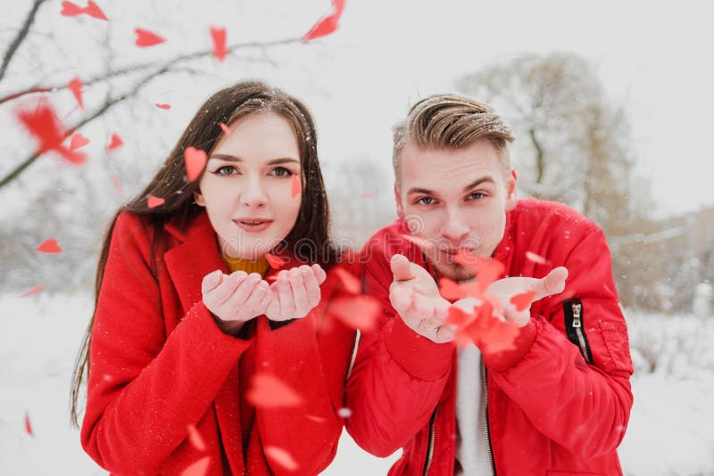 A young couple, a guy and a girl, on the street in winter in red clothes blowing on confetti in the form of hearts. Celebration, birthday, Valentine`s day. A young couple, a guy and a girl, on the street in winter in red clothes blowing on confetti in the form of hearts. Celebration, birthday, Valentine`s day.