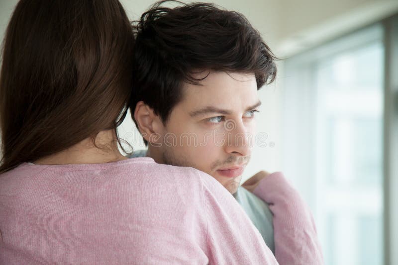 Woman hugging her upset boyfriend, sad guy looking away through the window, loving wife supporting husband, embracing and comforting him, helping to solve business problems, couple reconciliation. Woman hugging her upset boyfriend, sad guy looking away through the window, loving wife supporting husband, embracing and comforting him, helping to solve business problems, couple reconciliation