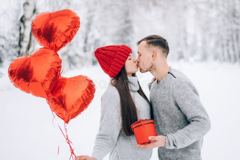 Young couple in love in the park on a background of snow and red heart-shaped balloons. Valentine`s day gift. The woman sits on