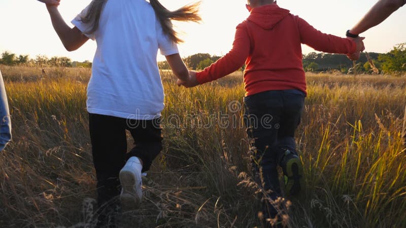 Young couple of parents with kids holding hands of each other and running through grass field at sunset. Mom and dad