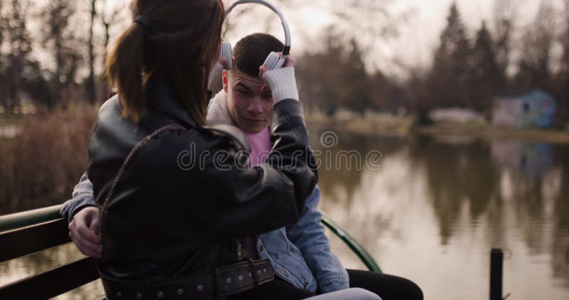 Young Couple partage son casque et aime la musique ensemble sur un banc de parc au bord du lac banque de vidéos