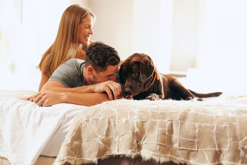 Shot of a young couple relaxing on the bed with her pet dog. Young men and women in bedroom playing with their dog in morning.