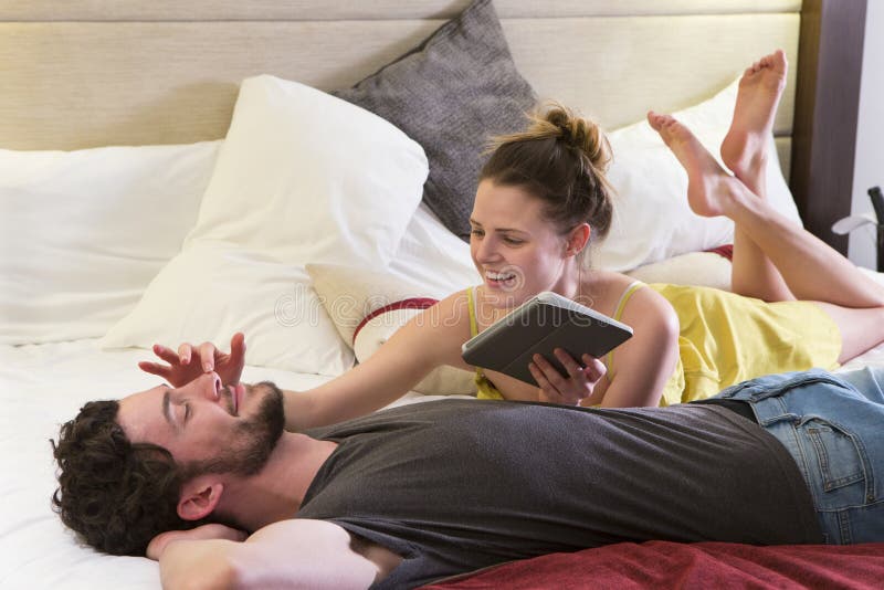 Young couple lying on their bed in their hotel room. The women is using a digital tablet and stroking her partners nose, who is lying on his back. Young couple lying on their bed in their hotel room. The women is using a digital tablet and stroking her partners nose, who is lying on his back.