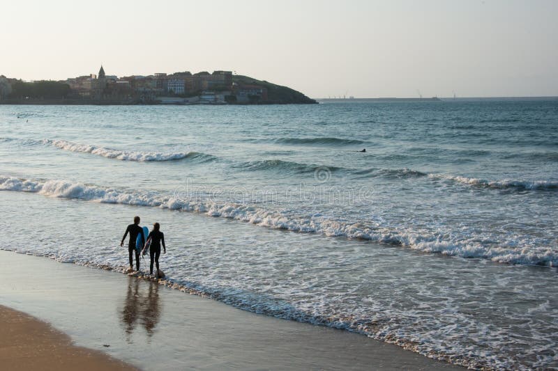 Young couple seen from their back entering the water to surf at Gijon beach at sunset. View of the city in the distance. Asturias, Spain, Europe