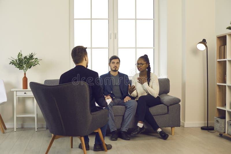 Couple experiencing relationship problem having therapy session at marriage and family therapist&#x27;s office. Young multiracial people trying to resolve conflict with help of professional psychologist. Couple experiencing relationship problem having therapy session at marriage and family therapist&#x27;s office. Young multiracial people trying to resolve conflict with help of professional psychologist