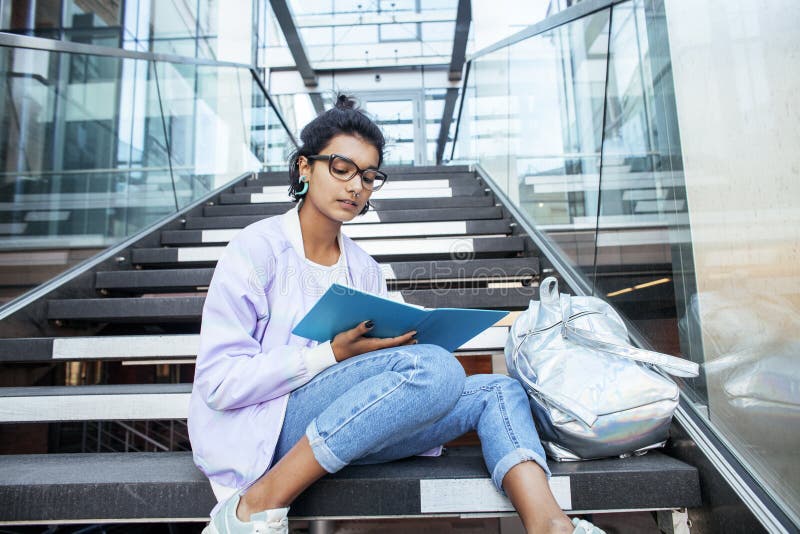 Young cute indian girl at university building sitting on stairs reading a book, wearing hipster glasses, lifestyle