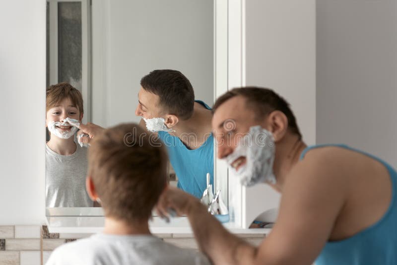 Young dad and his son in shaving foam on faces have fun in bathroom. Morning facial hygiene, time together