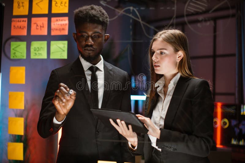 Technology, software programming, idea, people concept. Waist up portrait of two young diverse multiethnic coworkers, standing near the glass board, writing and using tablet pc