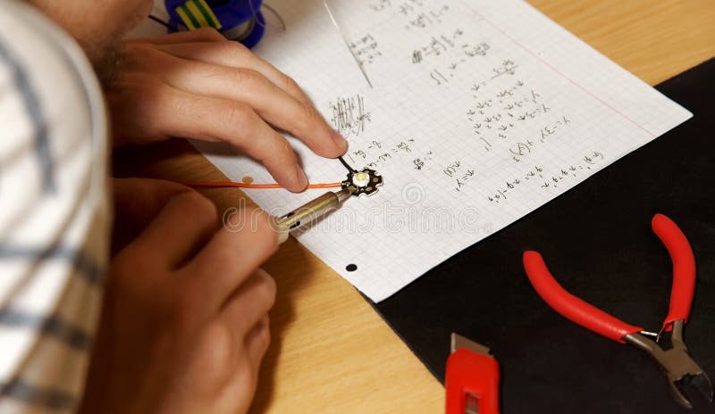 Young engineering college student soldering simple wired lambertian led diode on his desk, hands closeup, holding soldering iron. Temporary makeshift workspace table, mathematical notes, simple tools