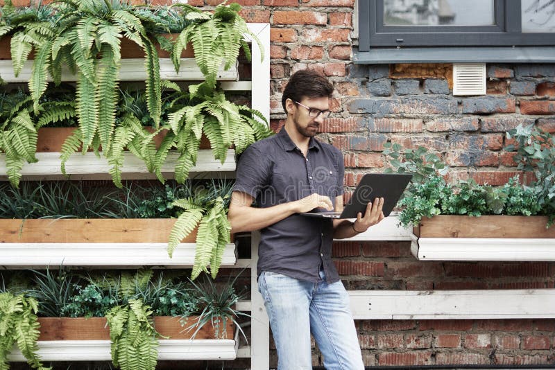 Young entrepreneur working outside using modern laptop. Standing near brick wall, plants, eco office. Successful business people royalty free stock image Young entrepreneur working outside using modern laptop. Standing near brick wall, plants, eco office. Successful business people royalty free stock image