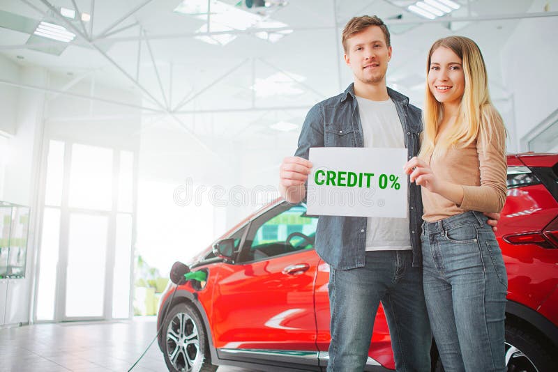 Young family buying first electric car in the showroom. Smiling attractive couple holding paper with word Credit while stock image Young family buying first electric car in the showroom. Smiling attractive couple holding paper with word Credit while stock image