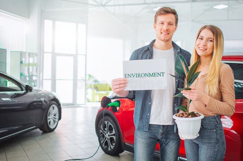 Young family buying first electric car in the showroom. Smiling attractive couple holding paper with word Environment royalty free stock photography Young family buying first electric car in the showroom. Smiling attractive couple holding paper with word Environment royalty free stock photography