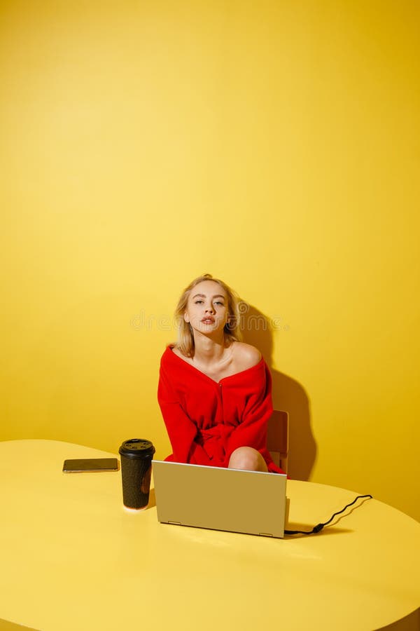 Young fashion girl blogger dressed in stylish red coat sitting at the yellow table with the laptop and cup of coffee on stock image Young fashion girl blogger dressed in stylish red coat sitting at the yellow table with the laptop and cup of coffee on stock image