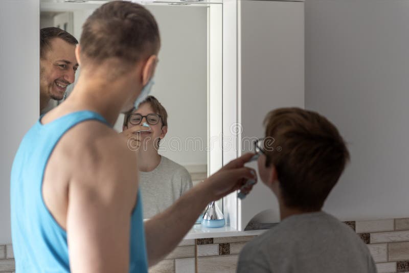 Young father and his teenage son with shaving foam on his nose. Dad and son in bathroom in front of mirror together