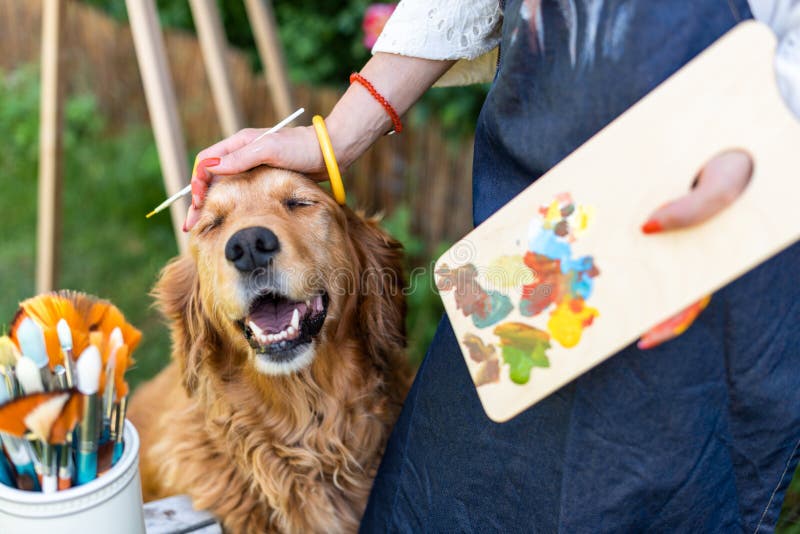 Young female artist working on her art canvas painting outdoors in her garden with golden retriever keeping her company.