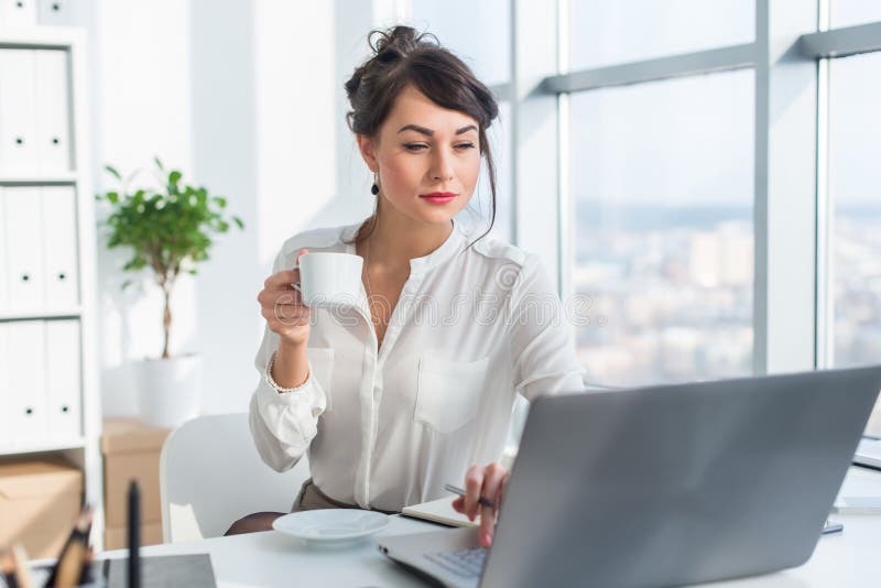 Young female business person working in office using laptop, reading and searching information attentively, drinking royalty free stock image Young female business person working in office using laptop, reading and searching information attentively, drinking royalty free stock image