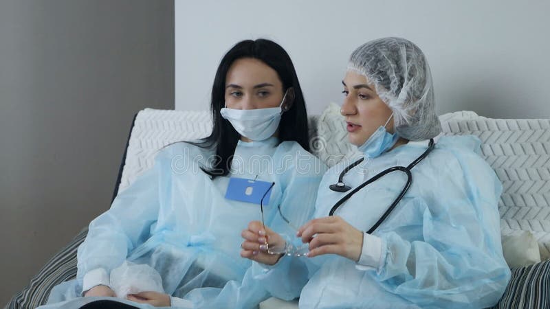 Young female doctors in blue uniform wearing mask sit and chat on sofa after hard work