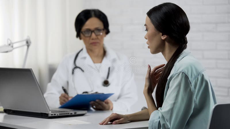 Young female patient saying symptoms, experienced doctor keeping medical records