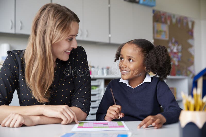 Young female primary school teacher working one on one with a schoolgirl at a table in a classroom, both looking at each other smiling, close up