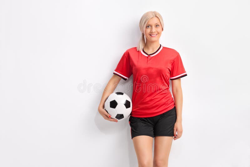 Young female soccer player in a red jersey holding a ball and leaning against a wall