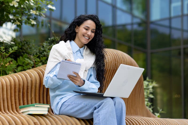 Young female student studying outside university campus, Latin American woman with laptop sitting on bench and recording class lecture, happy smiling.