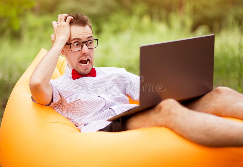 Young freelancer man with shocked face in shirt, red bow tie and glasses sitting on inflatable sofa with laptop in summer park