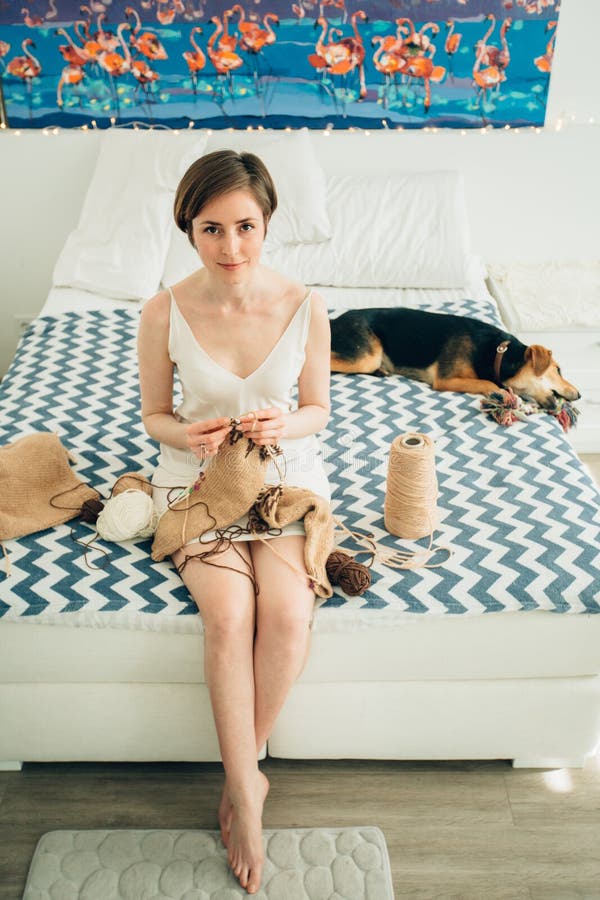 Young friendly girl craftwoman in nightdress looking at camera while knitting sweater on bed. Cute cur dog besides. Home, royalty free stock images Young friendly girl craftwoman in nightdress looking at camera while knitting sweater on bed. Cute cur dog besides. Home, royalty free stock images