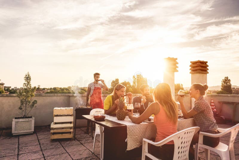 Young friends having barbecue party at sunset on penthouse patio - Happy people doing bbq dinner outdoor cooking meat and drinking