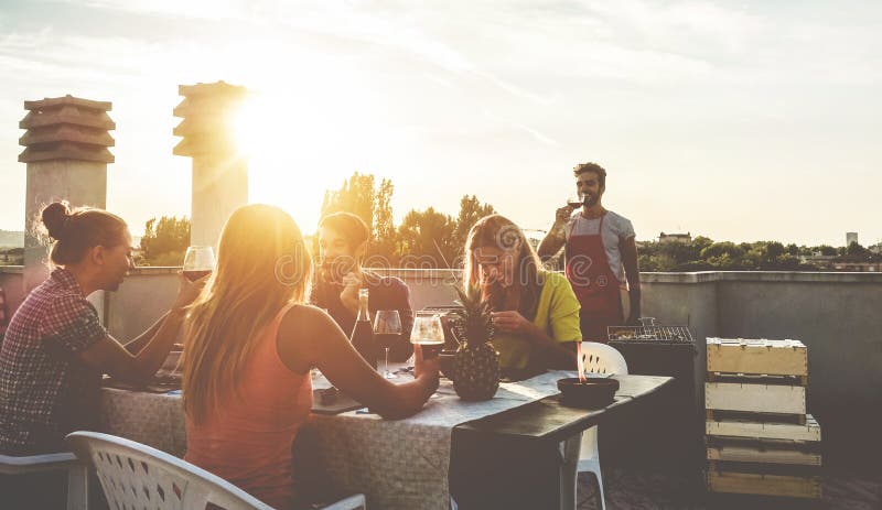 Young friends having barbecue party at sunset on penthouse patio - Happy people doing bbq dinner outdoor cooking meat and drinking