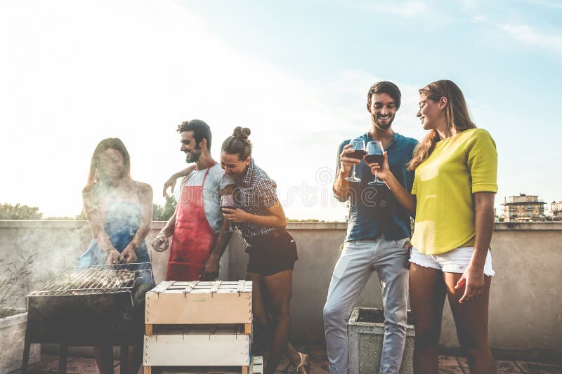 Young friends having barbecue party at sunset on terrace - Happy people doing bbq dinner outdoor cooking meat and drinking wine -